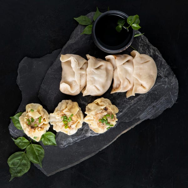 Filipino dumplings and siomai with green garnish and a small bowl of dark sauce on a slate board.